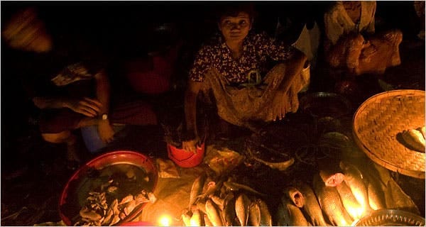 (ရန်ကုန်တွင် မီးပြတ်​နေသဖြင့်ဖ​ယောင်တိုင်ထွန်း၍ ​ဈေး​ရောင်​နေသူတစ်ဦး ။ဓါတ်ပုံ-The New Yourk Times)