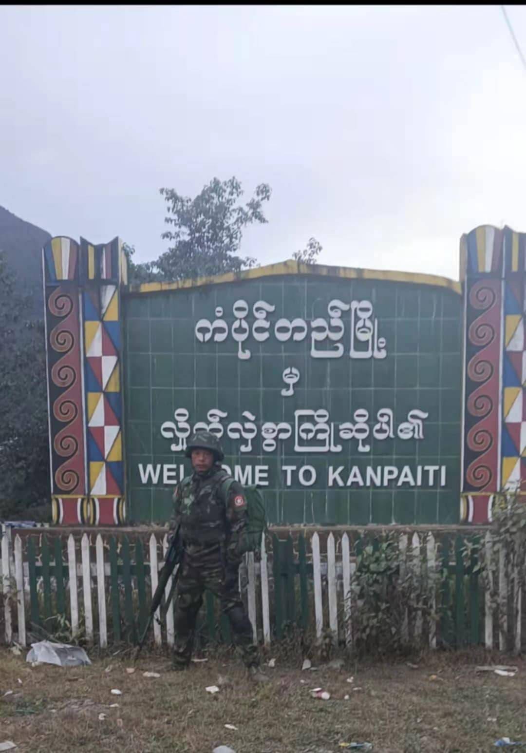 (ကံပိုက်တီ မြို့ဆိုင်းဘုတ် ​ရှေ့တွင် KIA တပ်ဖွဲ့ဝင်တဦး ။ဓါတ်ပုံ-​ပေးပို့)